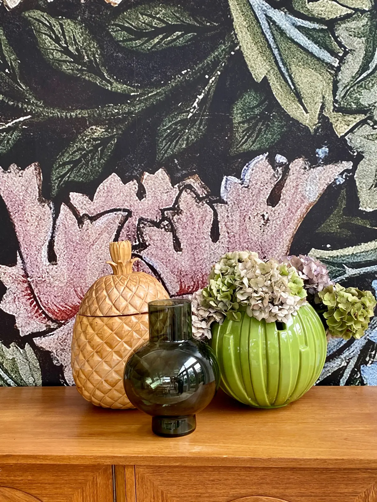 Green ceramic vases and hydrangeas on a sideboard against dark floral wallpaper