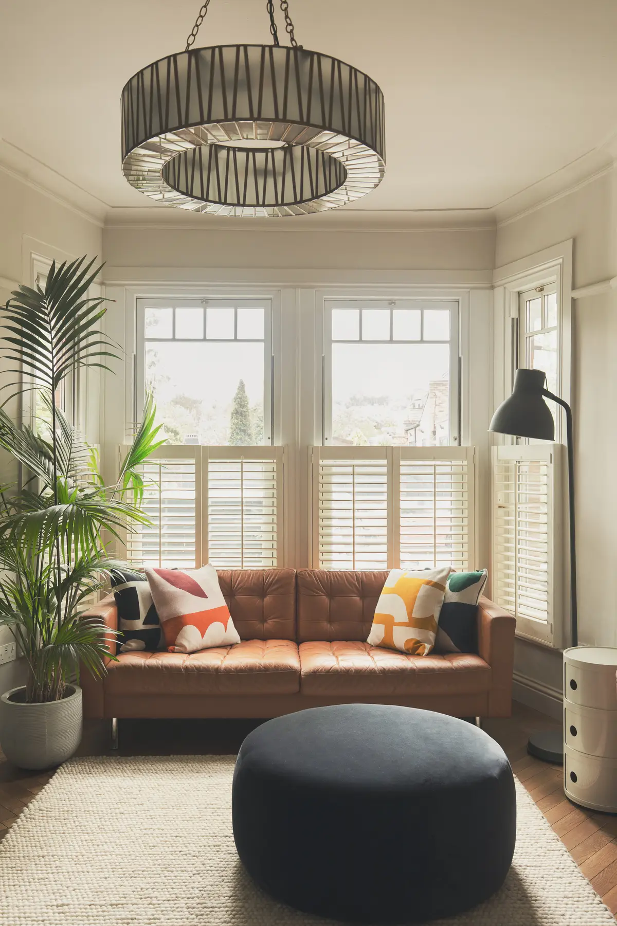Sunny living room with a tan leather sofa, patterned cushions and plantation shutters in a Crouch End home designed by JLV Design